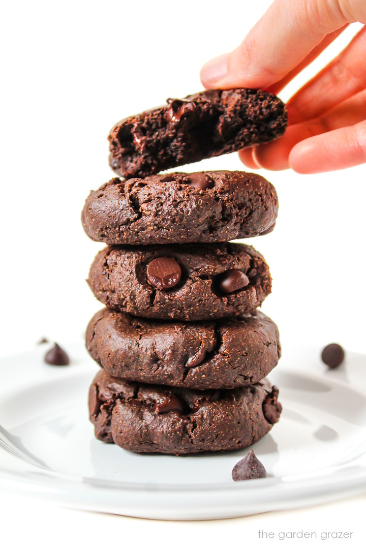 A hand taking the top vegan chocolate cookie from a stack on a white plate