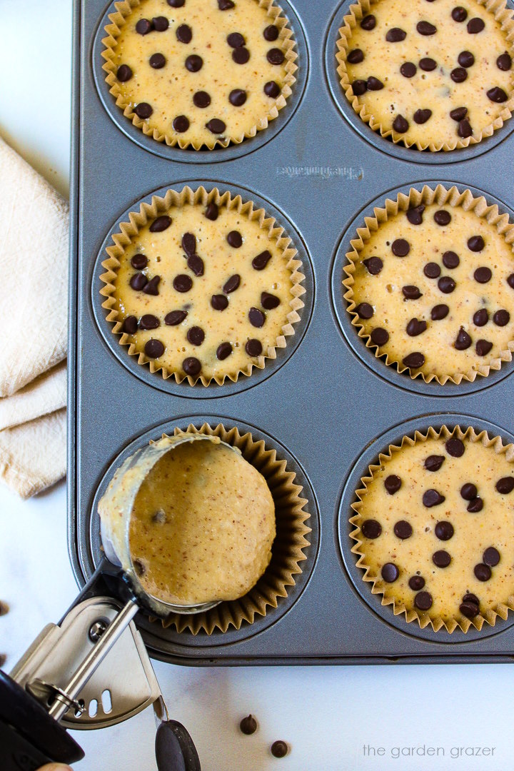An ice cream scoop with vegan chocolate chip muffin batter going into cupcake liners in a tray