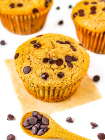 Three vegan chocolate chip muffins on a white table with chocolate chips scattered