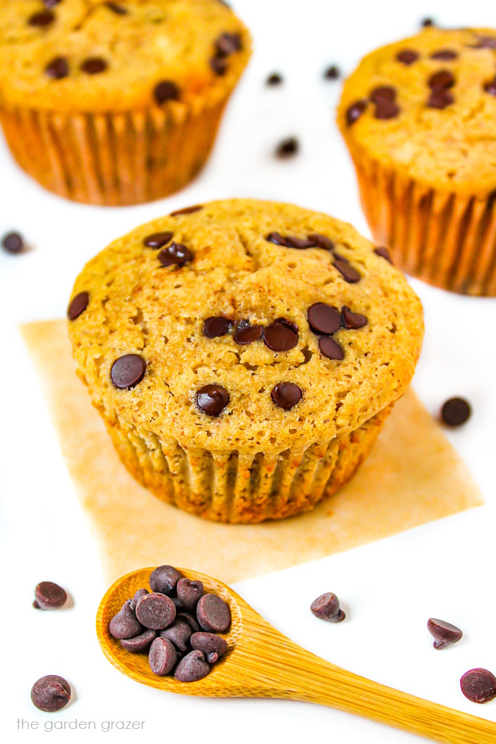 Three vegan chocolate chip muffins on a white table with chocolate chips scattered