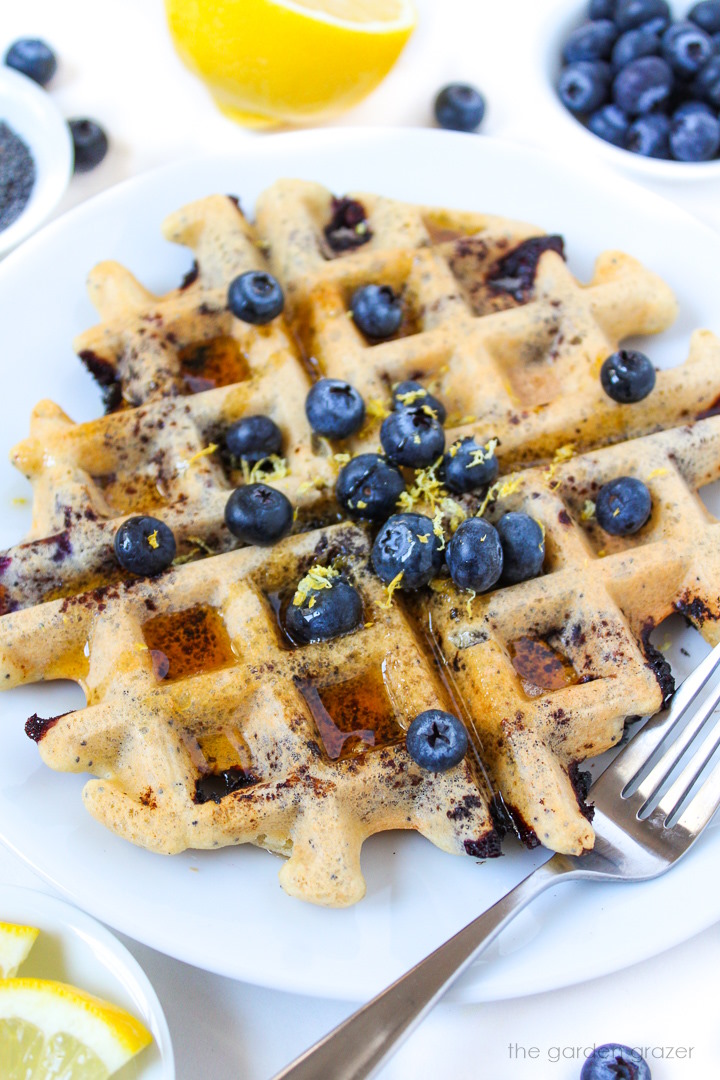 Blueberry waffles on a white plate topped with fresh berries, lemon zest, and maple syrup