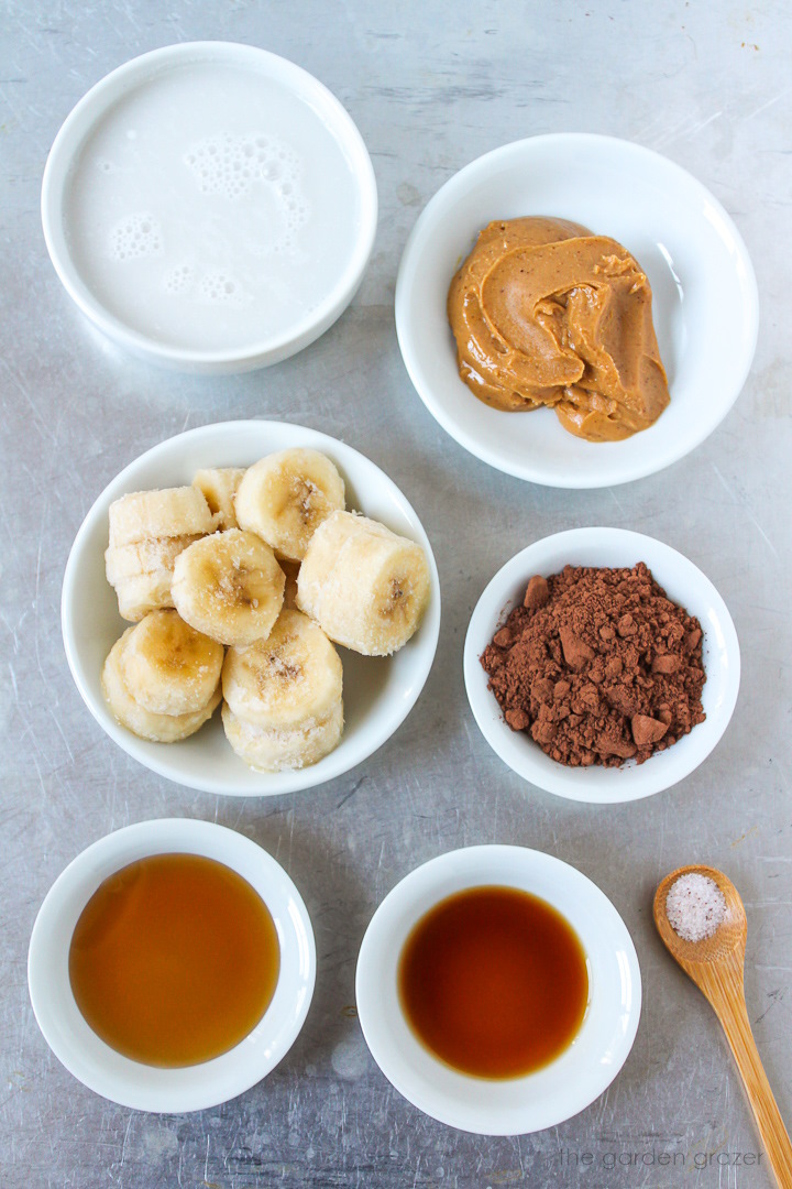 Sliced frozen banana, maple syrup, peanut butter, cocoa powder, dairy-free milk, and vanilla ingredients laid out on a metal tray