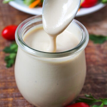 A spoon lifting up miso tahini dressing from a small glass jar on a wooden table