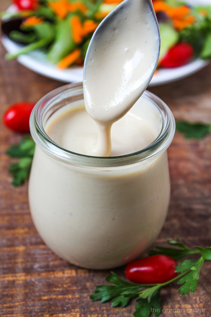 A spoon lifting up miso tahini dressing from a small glass jar on a wooden table