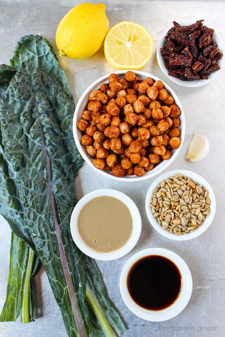 Fresh kale, sun-dried tomatoes, lemon, sunflower seeds, tahini, and garlic ingredients laid out on a metal tray