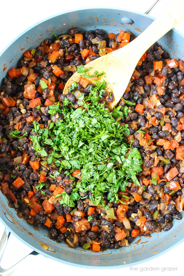 Overhead view of bean mixture cooking in a large skillet with wooden stirring spoon and fresh cilantro on top