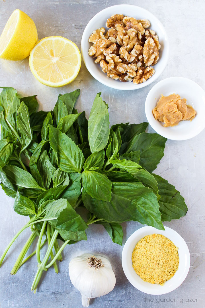 Fresh basil, nuts, lemon, garlic, miso, and nutritional yeast ingredients laid out on a metal tray