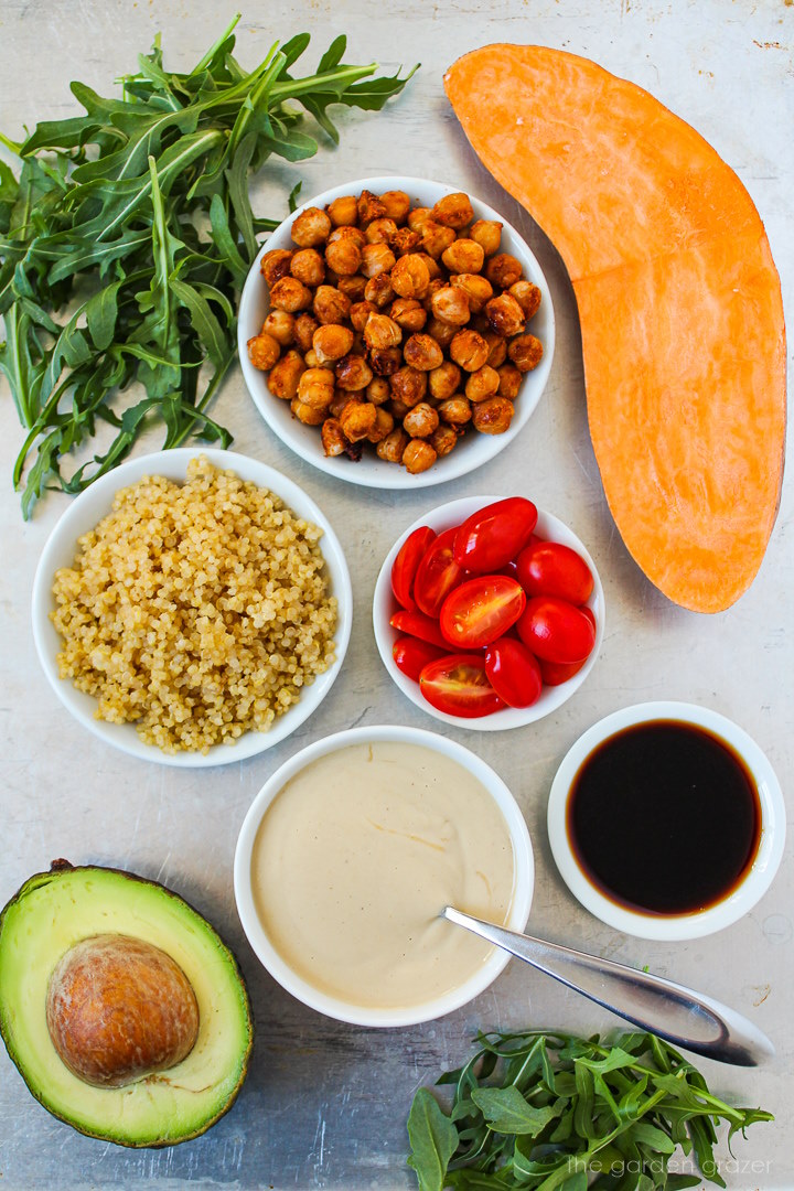 Fresh arugula, potato, roasted chickpeas, tomatoes, tahini dressing, and avocado ingredients laid out on a metal tray
