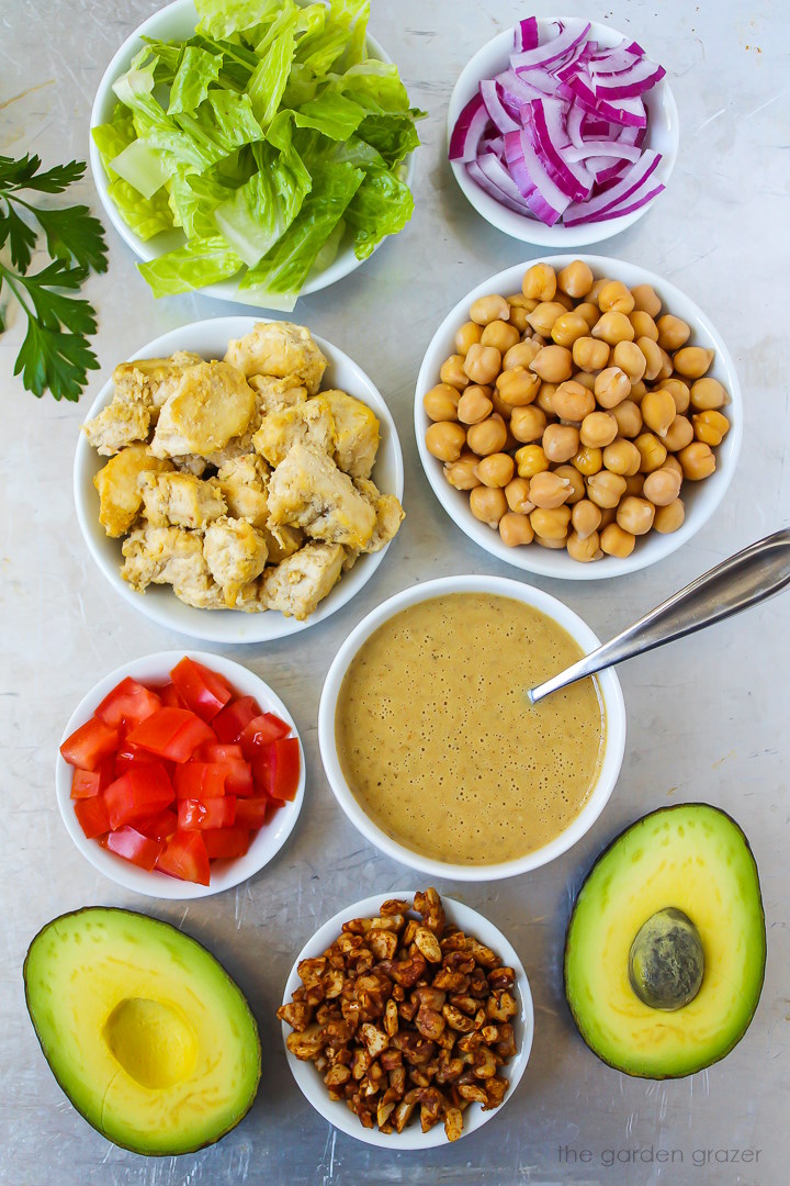 Chopped romaine, red onion, tomatoes, chickpeas, nut bacon, and dressing ingredients laid out on a metal tray