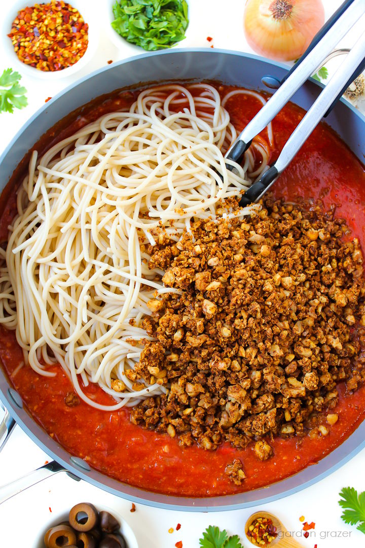Overhead view of preparing tomato sauce with noodles and chickpea-walnut crumbles in a large skillet before mixing together