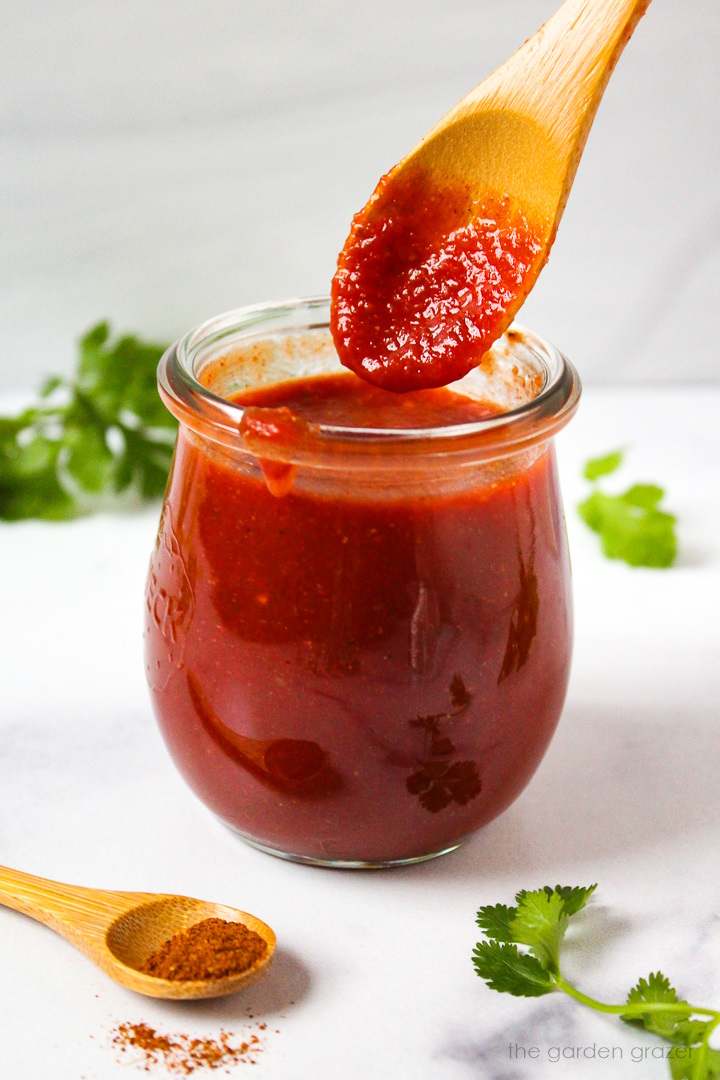 Serving spoon lifting up red taco sauce from a small glass jar