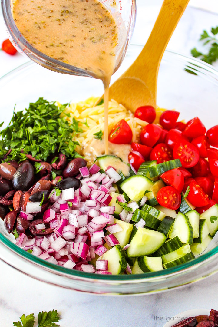 Pouring dressing over ingredients for Greek Orzo Pasta Salad before mixing together