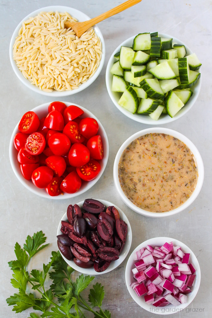 Overhead view of pasta, cucumber, grape tomato, olive, dressing, and red onion ingredients laid out on a metal tray