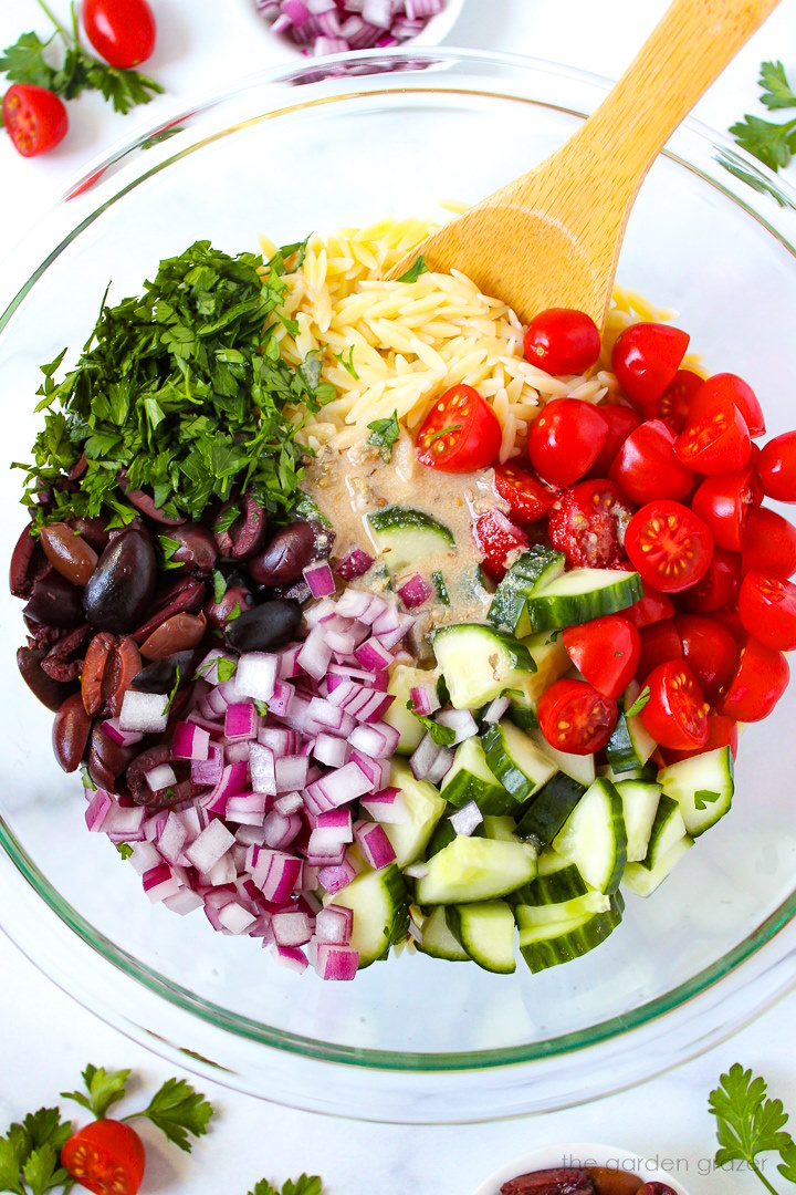 Overhead view of preparing Greek orzo pasta salad in a large glass bowl before mixing together