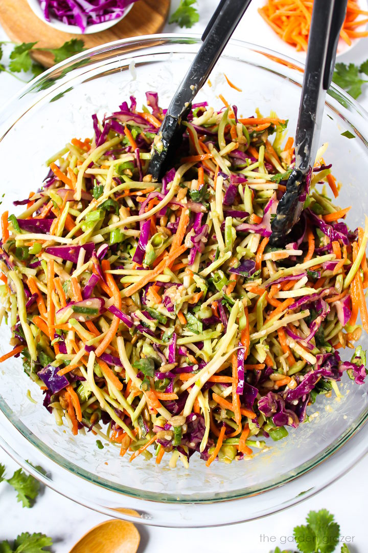 Asian broccoli slaw tossed with sesame ginger dressing in a glass mixing bowl with tongs
