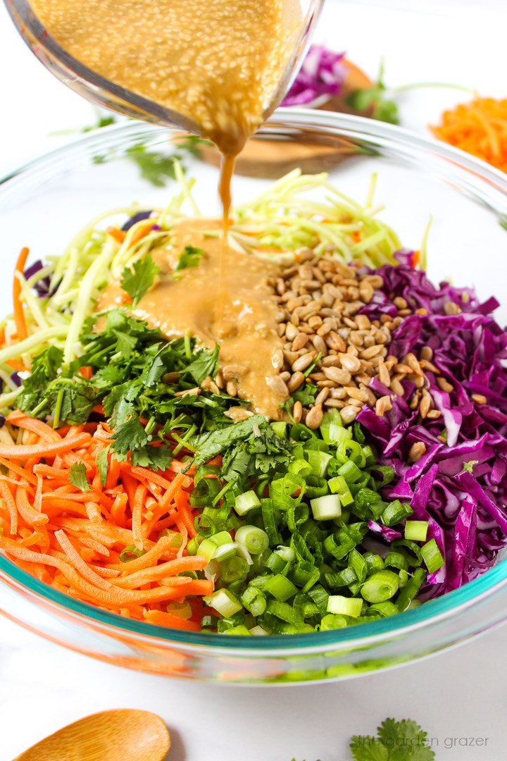 Sesame ginger dressing being poured over prepared broccoli slaw ingredients in a glass mixing bowl