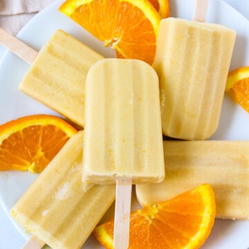Frozen vegan orange creamsicle popsicles arranged on a white serving place with fresh orange slices