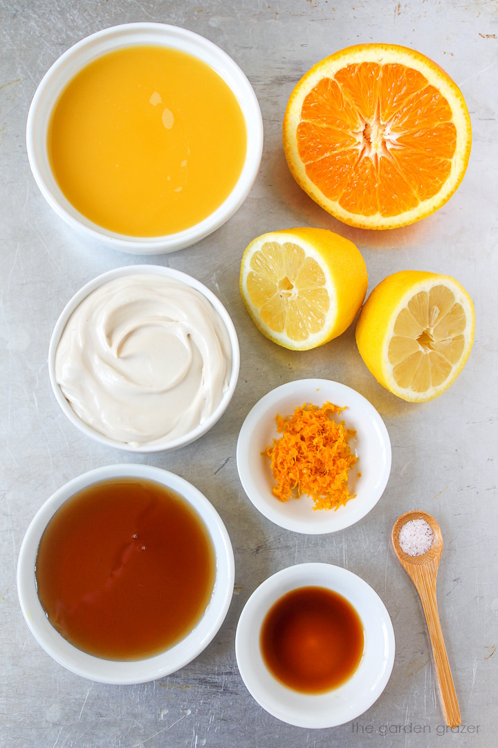 Orange juice, fresh oranges, lemons, Greek yogurt, orange zest, maple syrup, and vanilla ingredients laid out on a metal tray