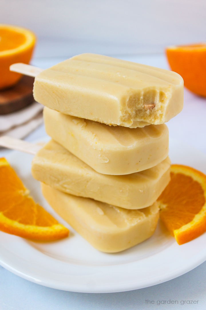 Frozen vegan orange creamsicle popsicles stacked on a white plate with fresh orange slices